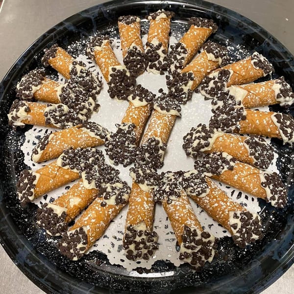 A platter of Italian cannolis, filled with cream, dipped in chocolate chips, and dusted with powdered sugar.
