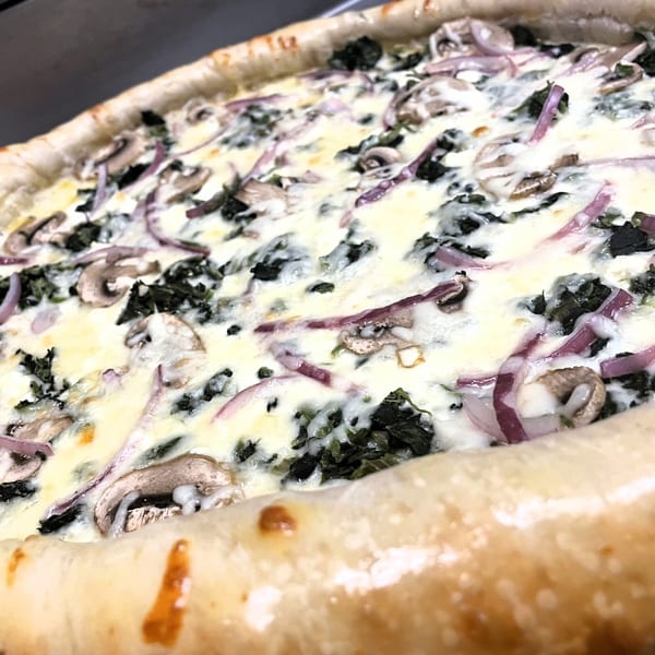 Close-up of a white pizza with spinach, mushrooms, and red onions, featuring a thick crust.