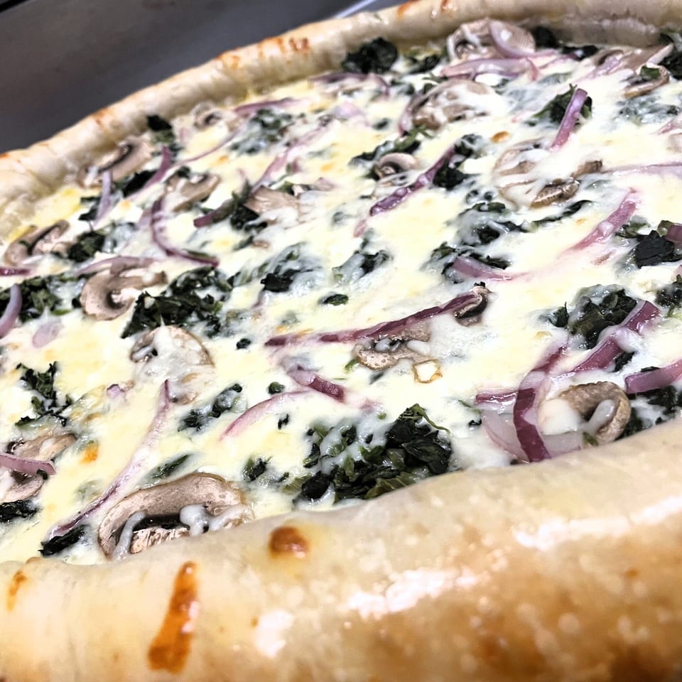 Close-up of a white pizza with spinach, mushrooms, and red onions, featuring a thick crust.