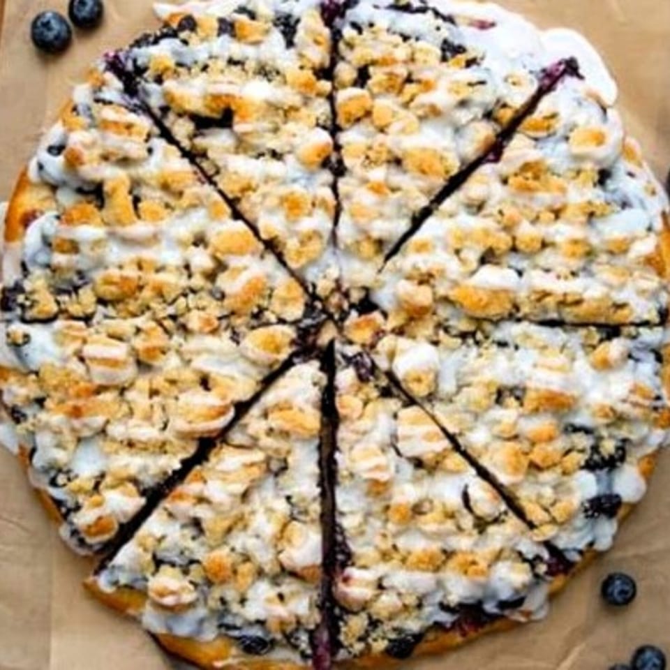 Blueberry dessert pizza with white icing and crumble topping, sliced into eight pieces.