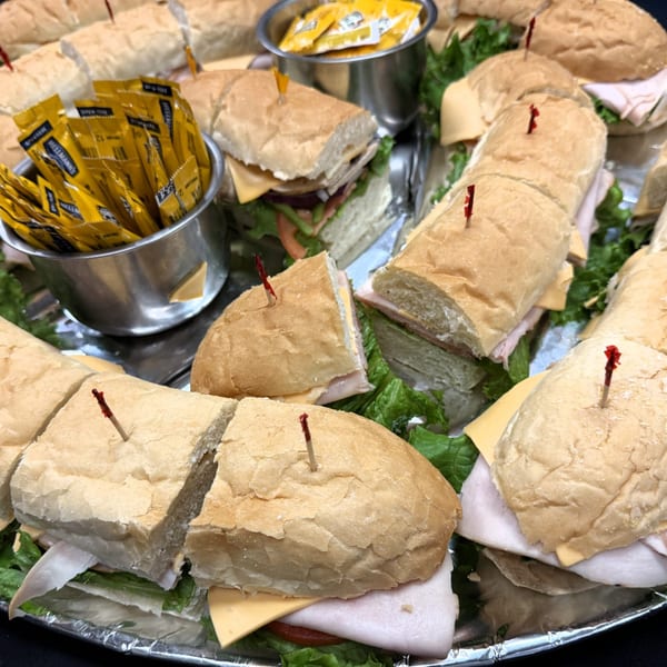A catering tray filled with sliced Italian sub sandwiches, lettuce, cheese, and condiment packets.