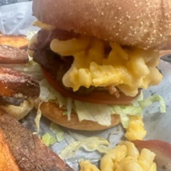 A burger topped with mac and cheese, lettuce, and tomato, served with potato wedges.