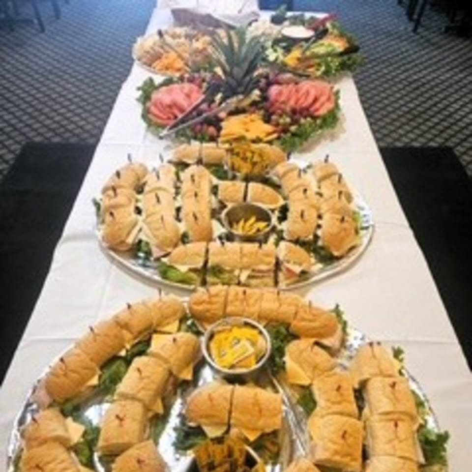 A long table laden with platters of Italian deli sandwiches, meats, cheeses, and fruit.